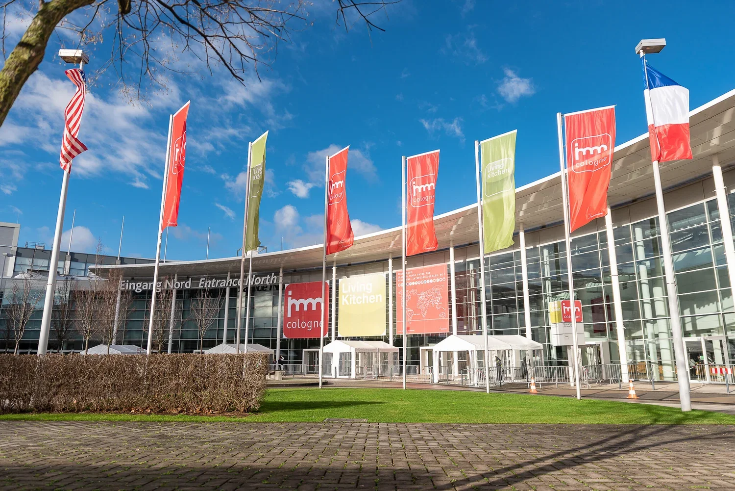Exterior view of the Cologne Exhibition Center with flags and banners, featuring the words 'IMM Cologne' and 'Living Kitchen' in front of a modern glass building under a partly cloudy sky.