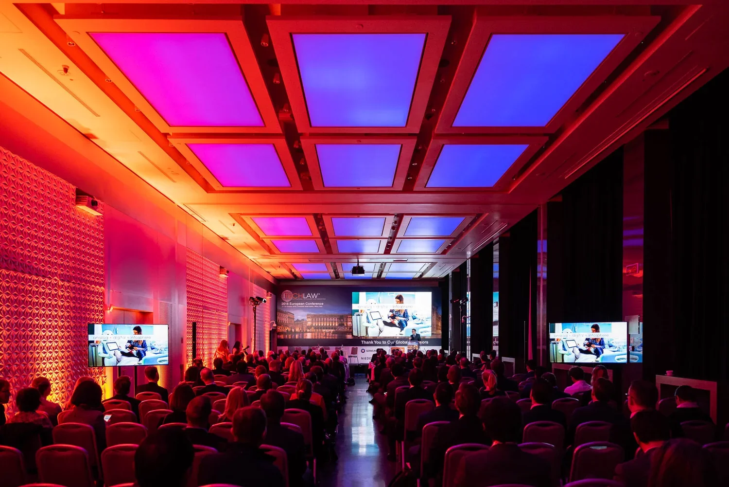 Conference room filled with attendees watching a presentation on stage, with large screens and colorful ceiling lighting.
