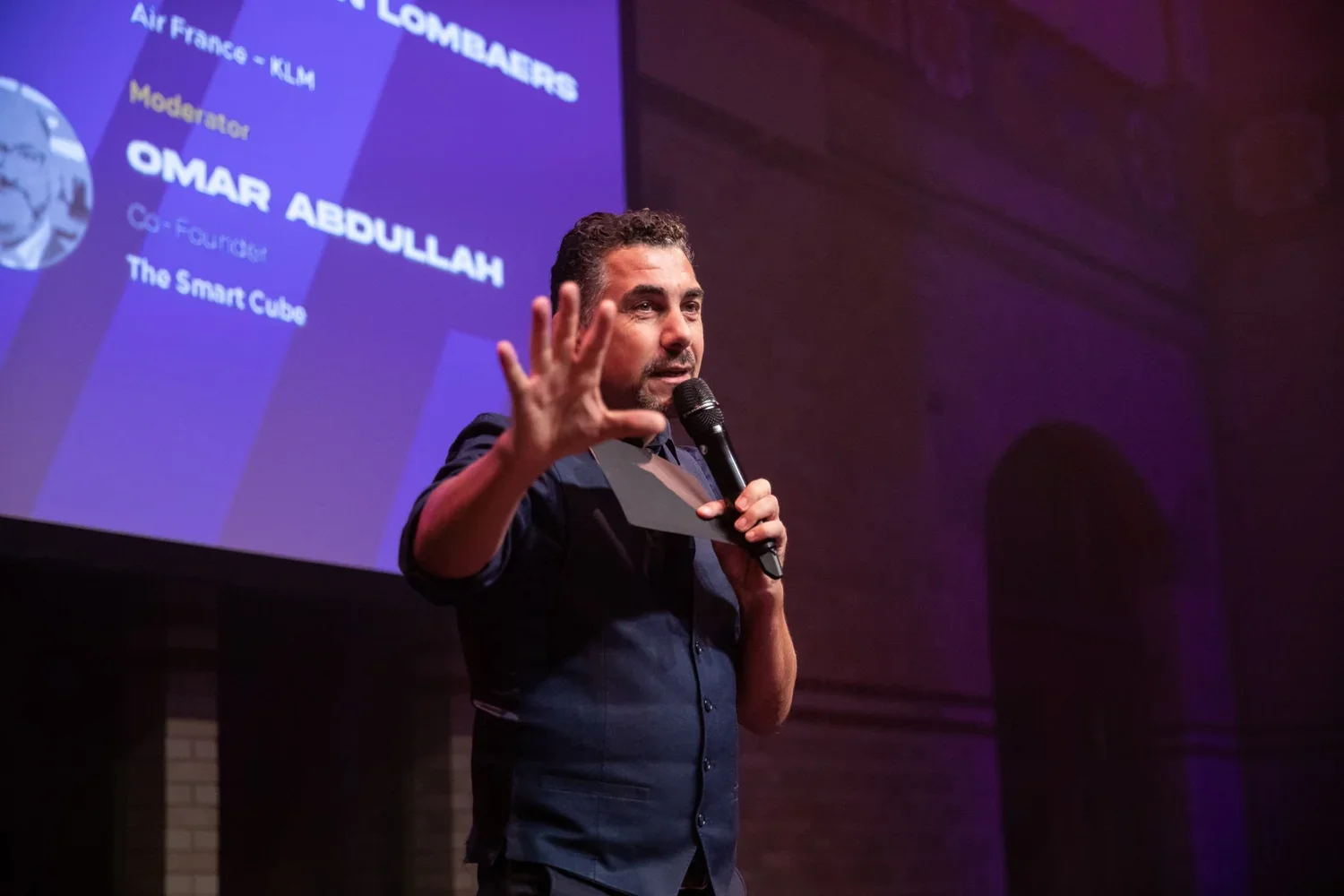 Man with short dark hair and beard speaking into a microphone on stage, gesturing with his left hand, with a purple presentation slide behind him displaying his name, Omar Abdallah, and titles, at an indoor event.