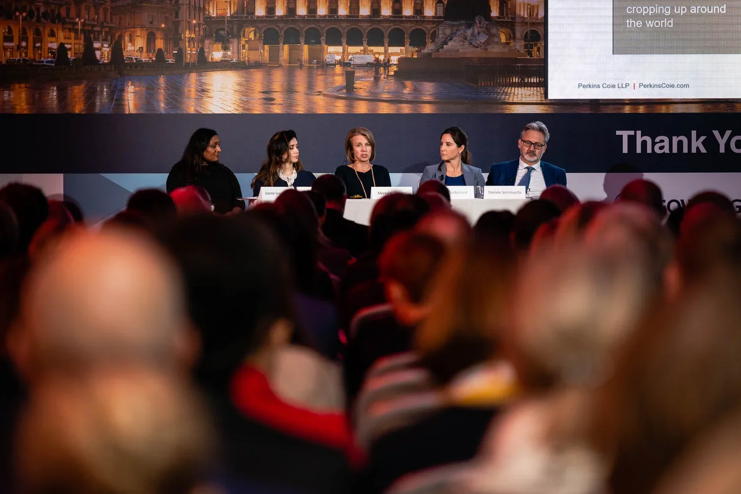 Panel of five people sitting at a table during a conference or panel discussion, with audience in foreground and a large cityscape image projected on the screen behind them.