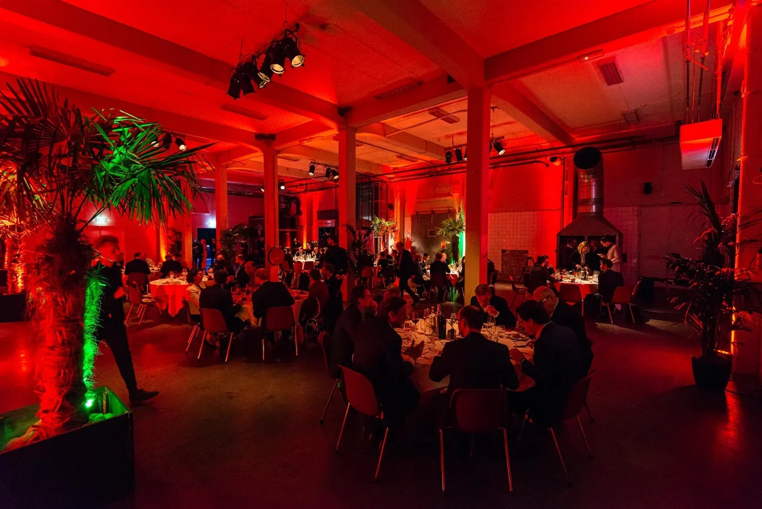 Guests dining at tables in a dimly lit, red-toned event hall with plants and decorative lighting.
