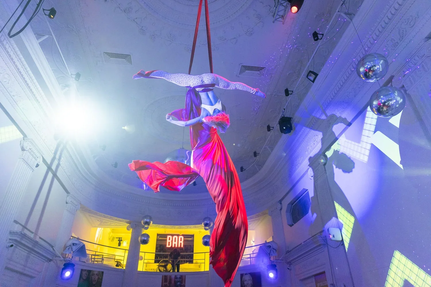 Aerial silk performers dancing in a grand, ornate hall with blue and purple lighting, mirrored disco balls, and a lit bar area below.