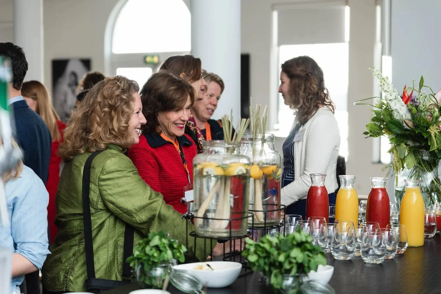 Attendees networking at corporate event drinks reception with colorful juices and floral table display — event photography Ballo Studio