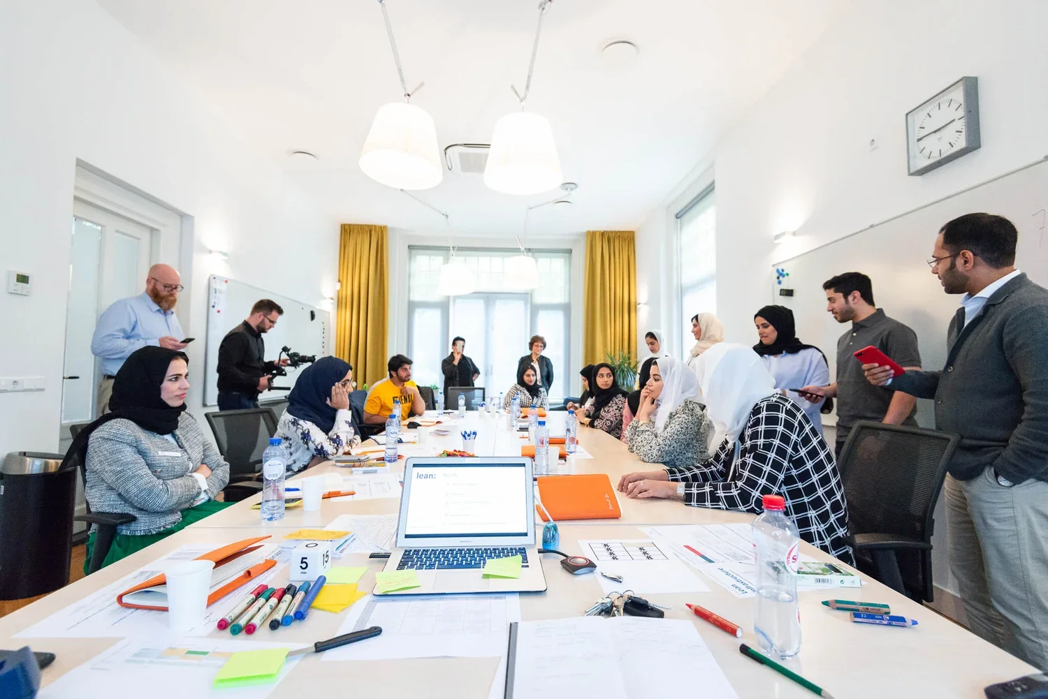 A diverse group of people seated around a large conference table in a bright room, with some standing and others listening or using their phones or tablets, while a few are speaking or presenting.