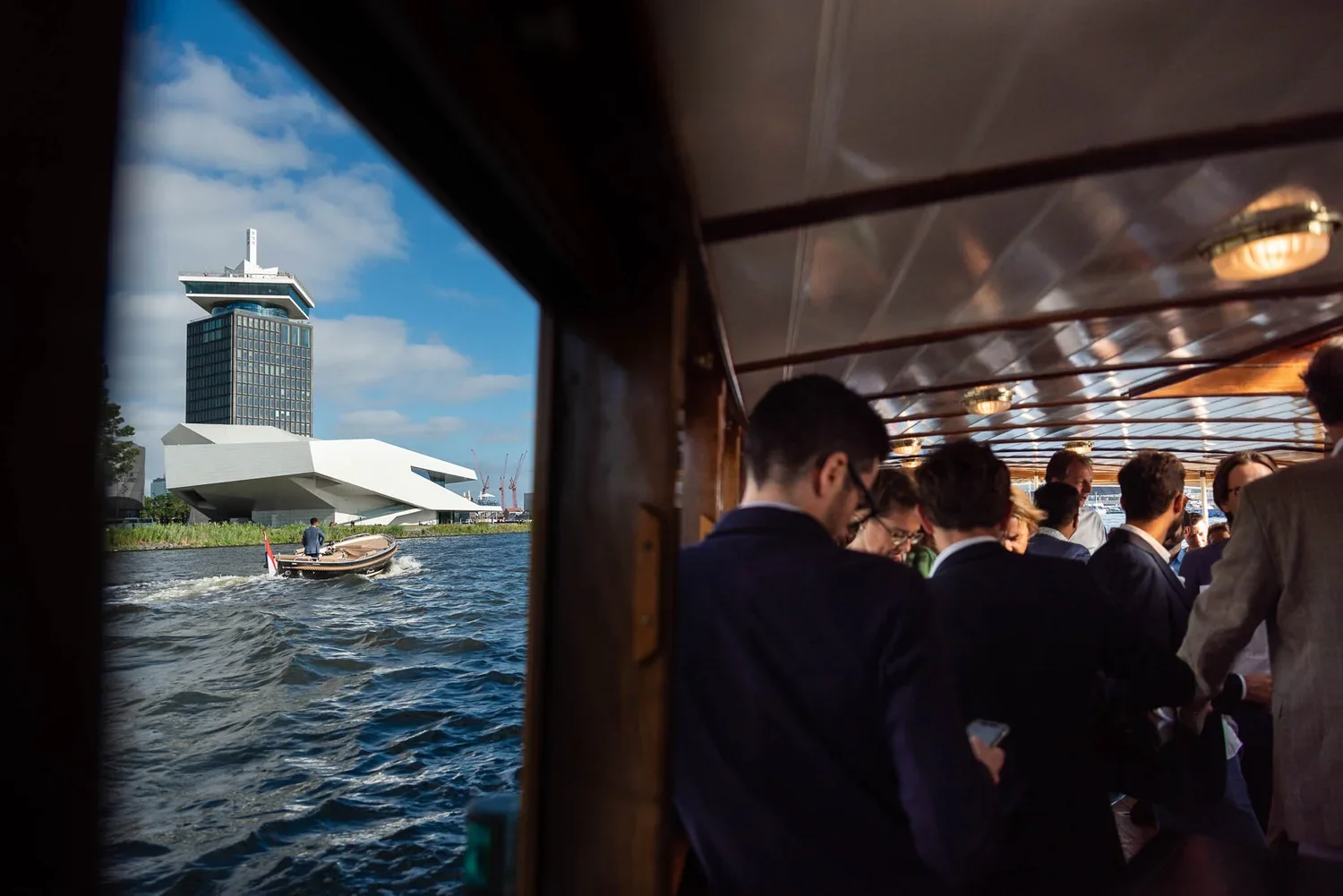 People in formal attire on a boat, water and modern buildings in the background, partly clear sky.