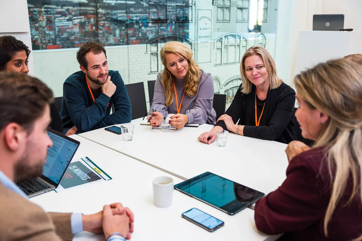 A diverse group of seven people sitting around a white conference table in a business meeting, with some using laptops and tablets, and a cityscape view through a large window in the background.