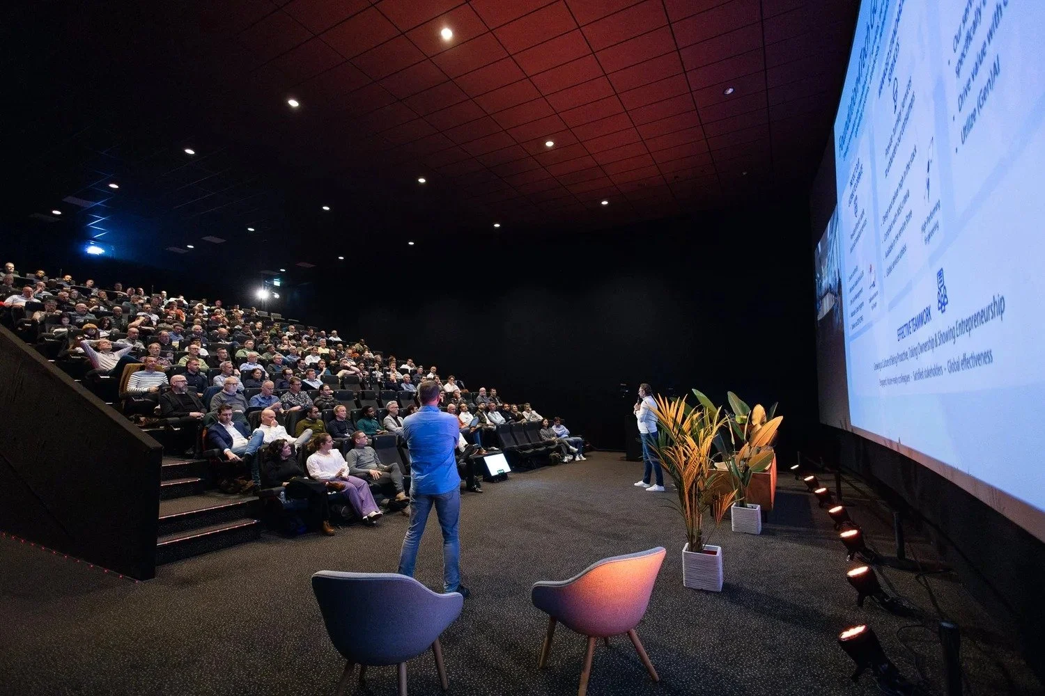 Speaker addressing full audience in modern cinema-style auditorium with tiered seating at corporate event Amsterdam — conference photography Amsterdam Ballo Studio