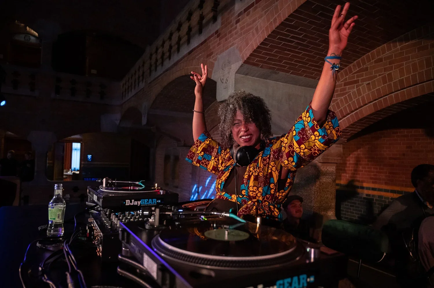A female DJ with curly hair and colorful patterned shirt, smiling and dancing with her hands raised while standing at her DJ equipment in a dimly lit venue.