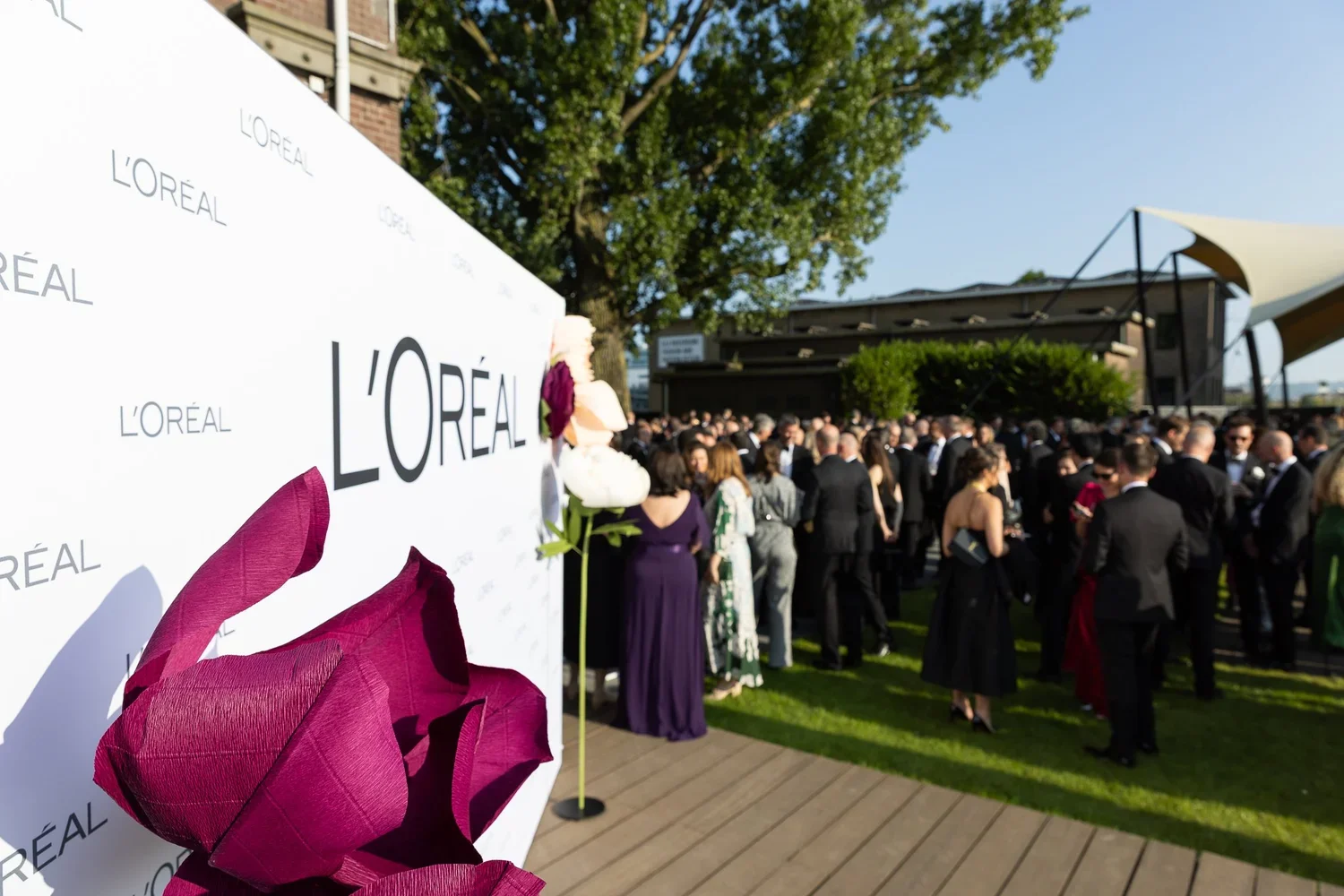 A crowd of people dressed in formal attire gathered outdoors near a white L'Oréal branded backdrop decorated with flowers for a daytime event.