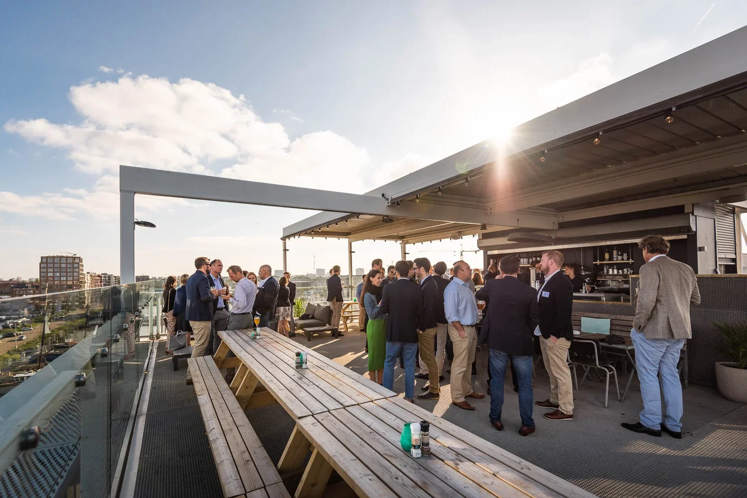 People socializing on a rooftop patio during a late afternoon or evening with the sun setting, skyline in the background, some sitting while others are standing near the bar.