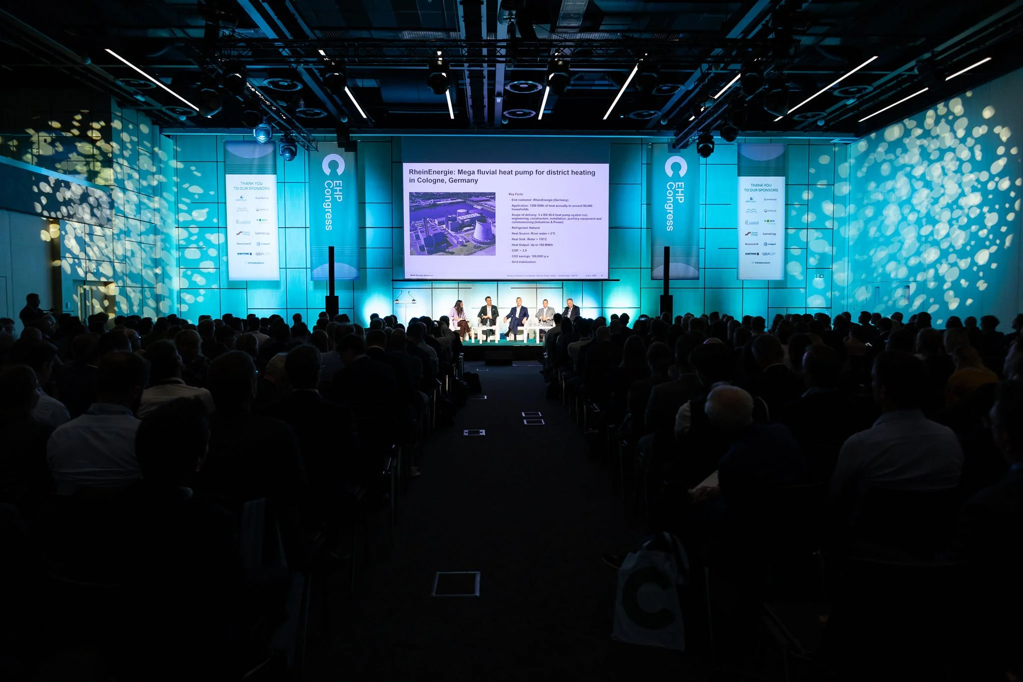 Conference with a large audience, panel of speakers on stage, large screen displaying a presentation about a heat pump in Cologne, Germany, with blue lighting and banners.