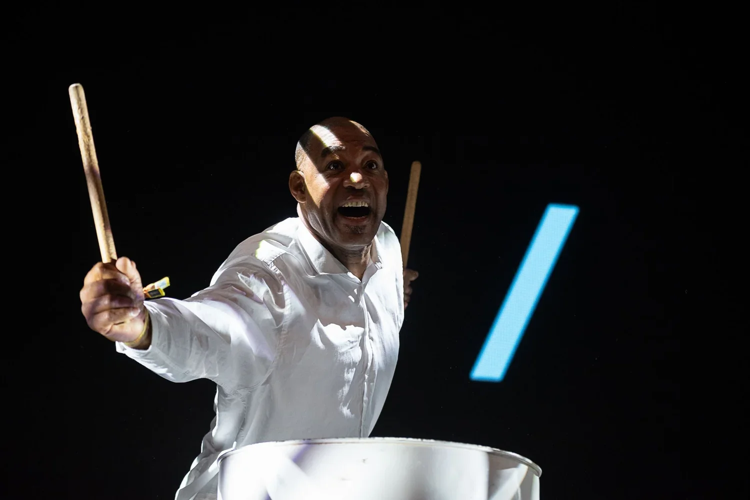 A man in a white shirt joyfully playing drums against a black background, with colorful lights.