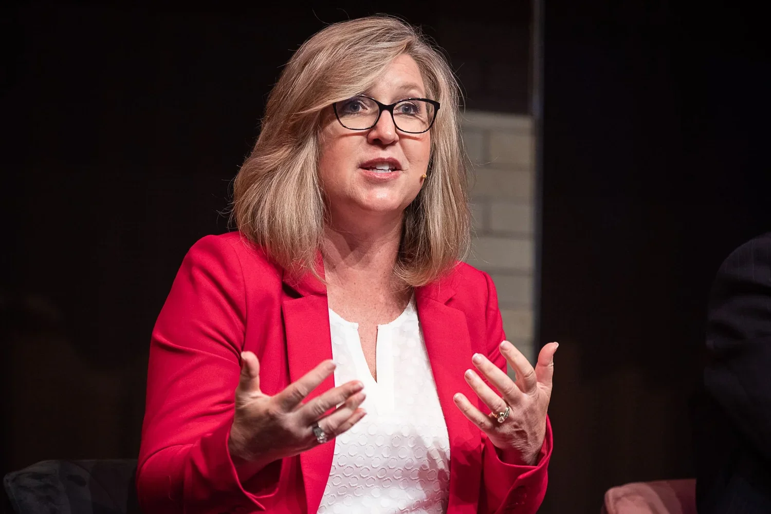 A woman with shoulder-length blonde hair, wearing glasses, a red blazer, and a white top, speaking or explaining with her hands.