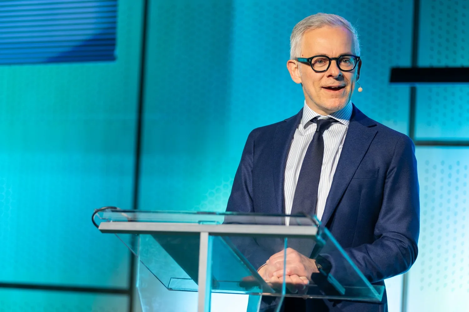 A man with gray hair, black glasses, wearing a dark blue suit, white and blue striped shirt, and striped tie, stands behind a glass podium on a stage with a colorful, modern background.
