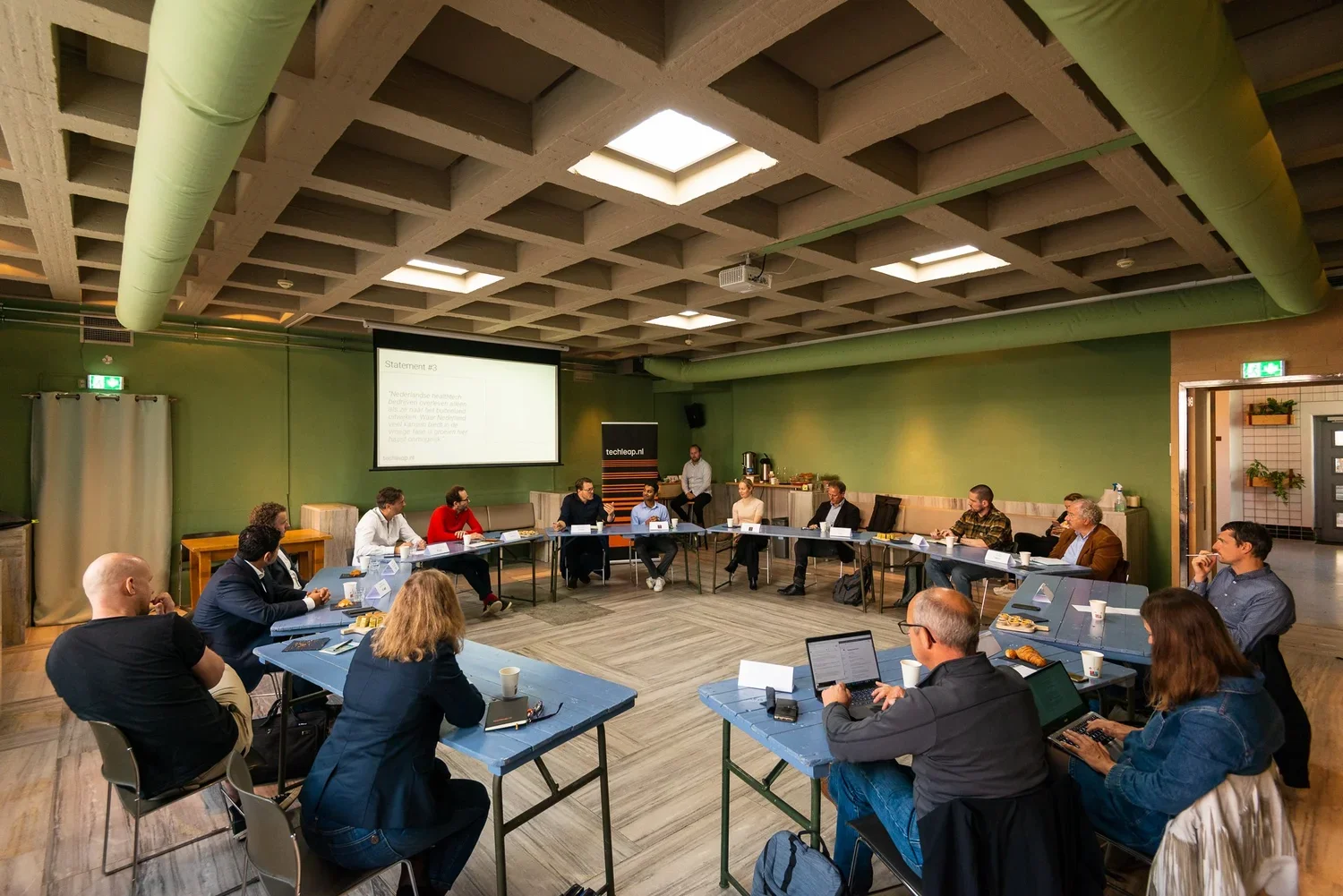 A group of people attending a meeting or conference in a room with green walls and a coffered ceiling. They are seated around a U-shaped arrangement of tables, with some looking at laptops and others listening to a presenter at the front. There is a 
