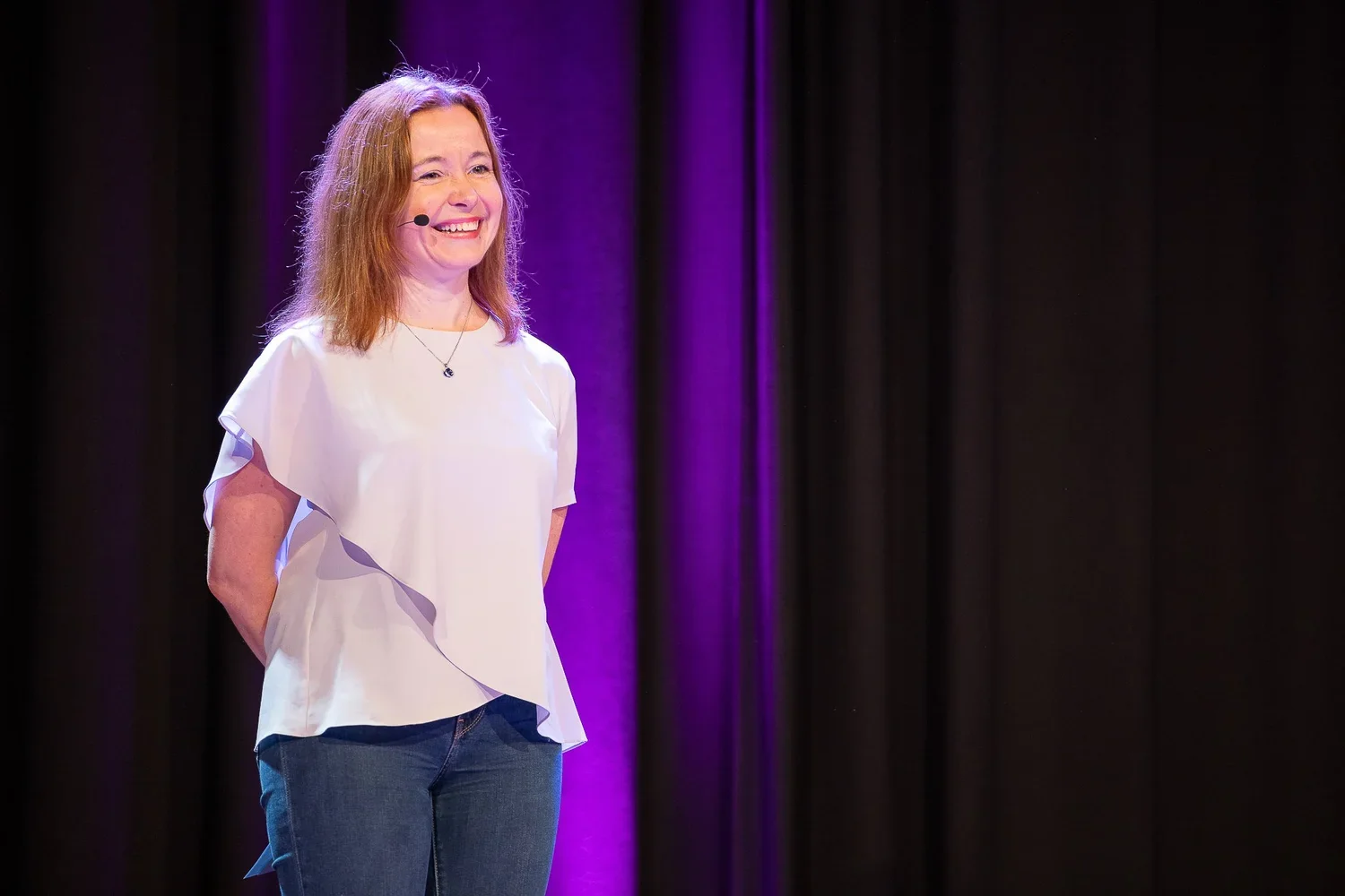 A woman with red hair smiling and standing on stage with purple lighting and black curtains in the background, wearing a white top and jeans.