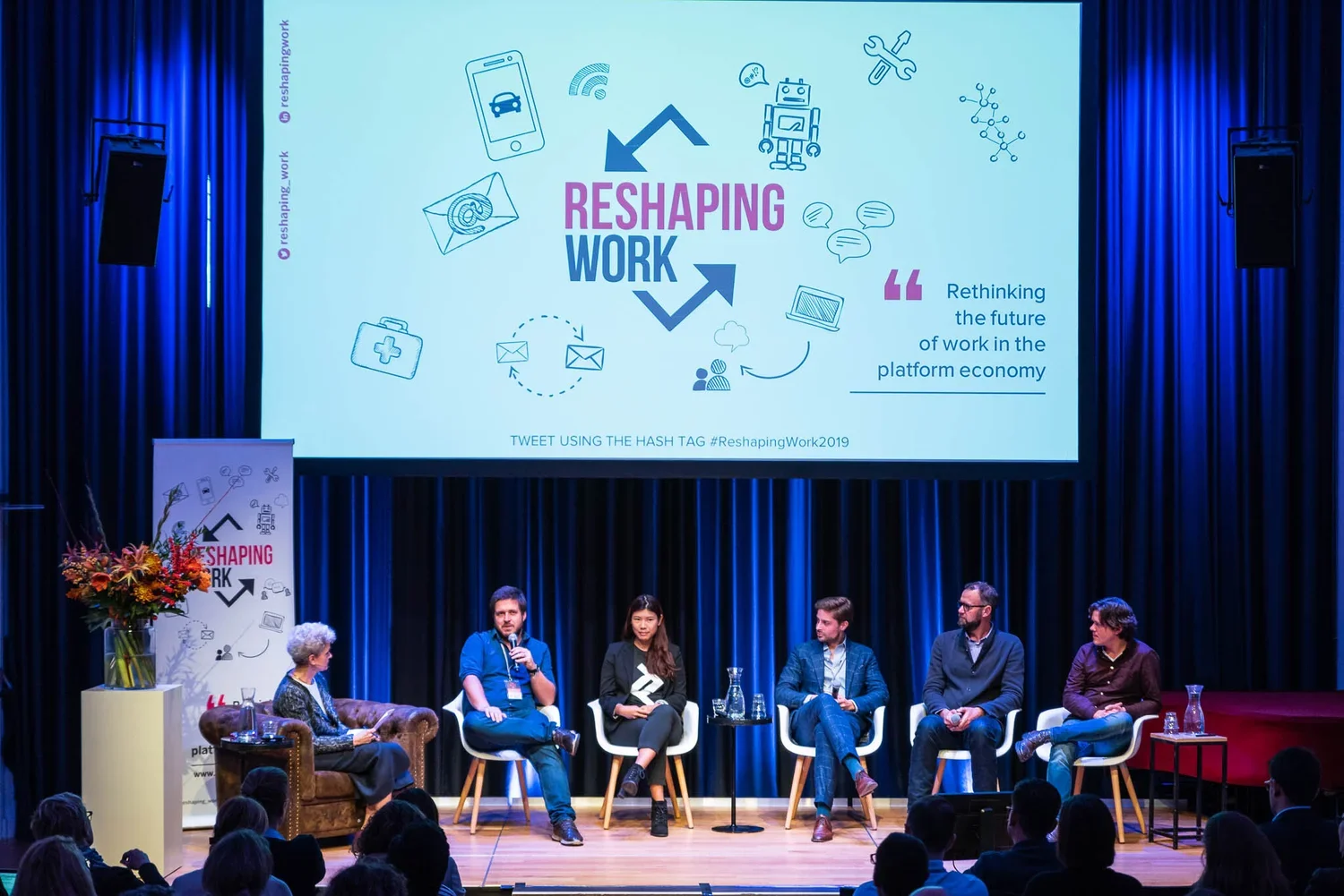 Panel discussion at a conference with a large screen displaying the event's theme 'Reshaping Work' and various related icons, including a robot, email, and a smartphone. Five speakers seated on stage, with a woman and four men, are engaging with the 