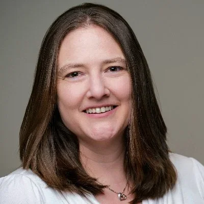 A woman with long brown hair smiling at the camera, wearing a white top and a necklace.