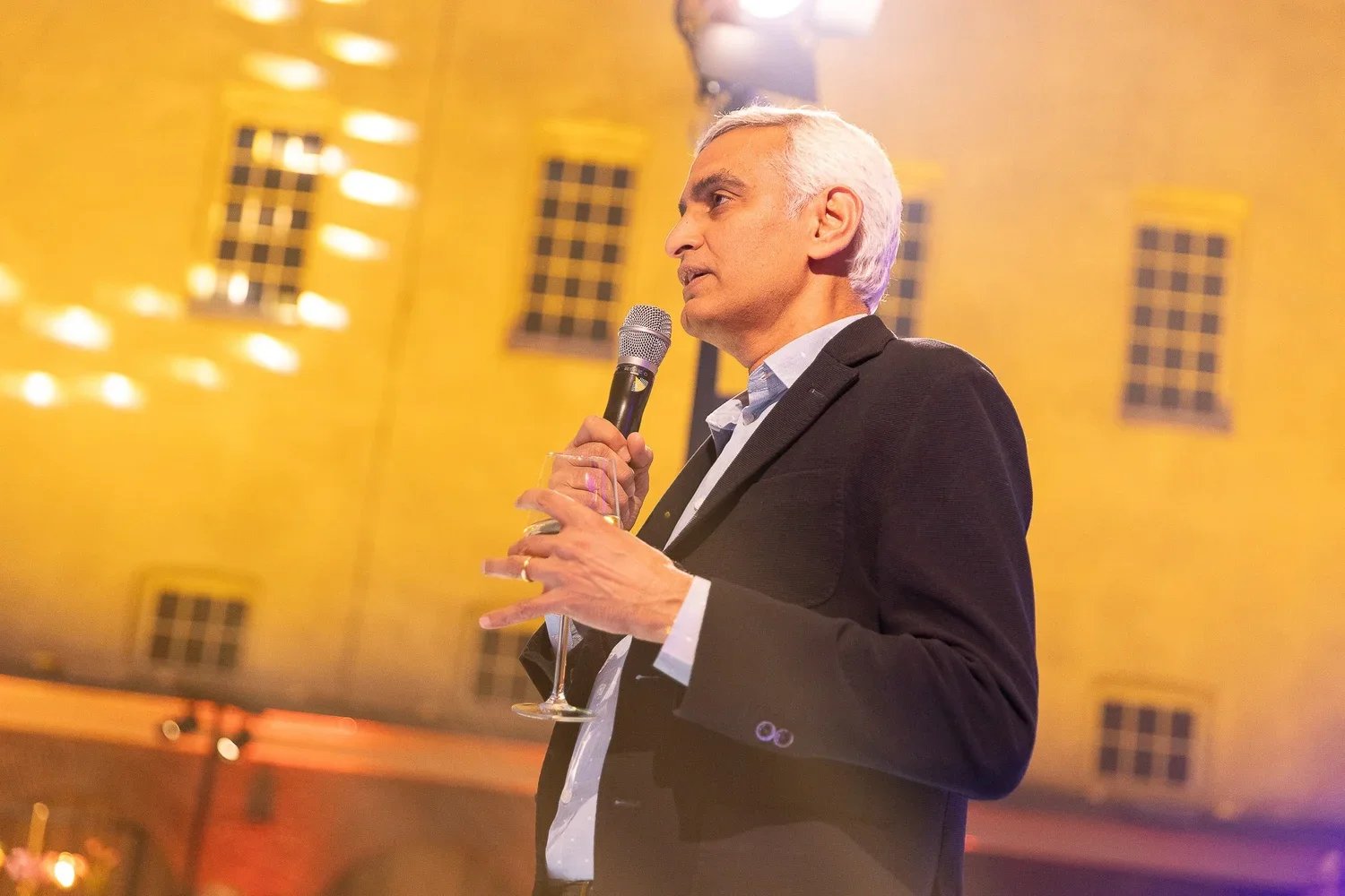 A man with gray hair wearing a black suit and white shirt, holding a microphone in one hand and a glass of drink in the other, speaking at an event in a room with yellow walls and windows.