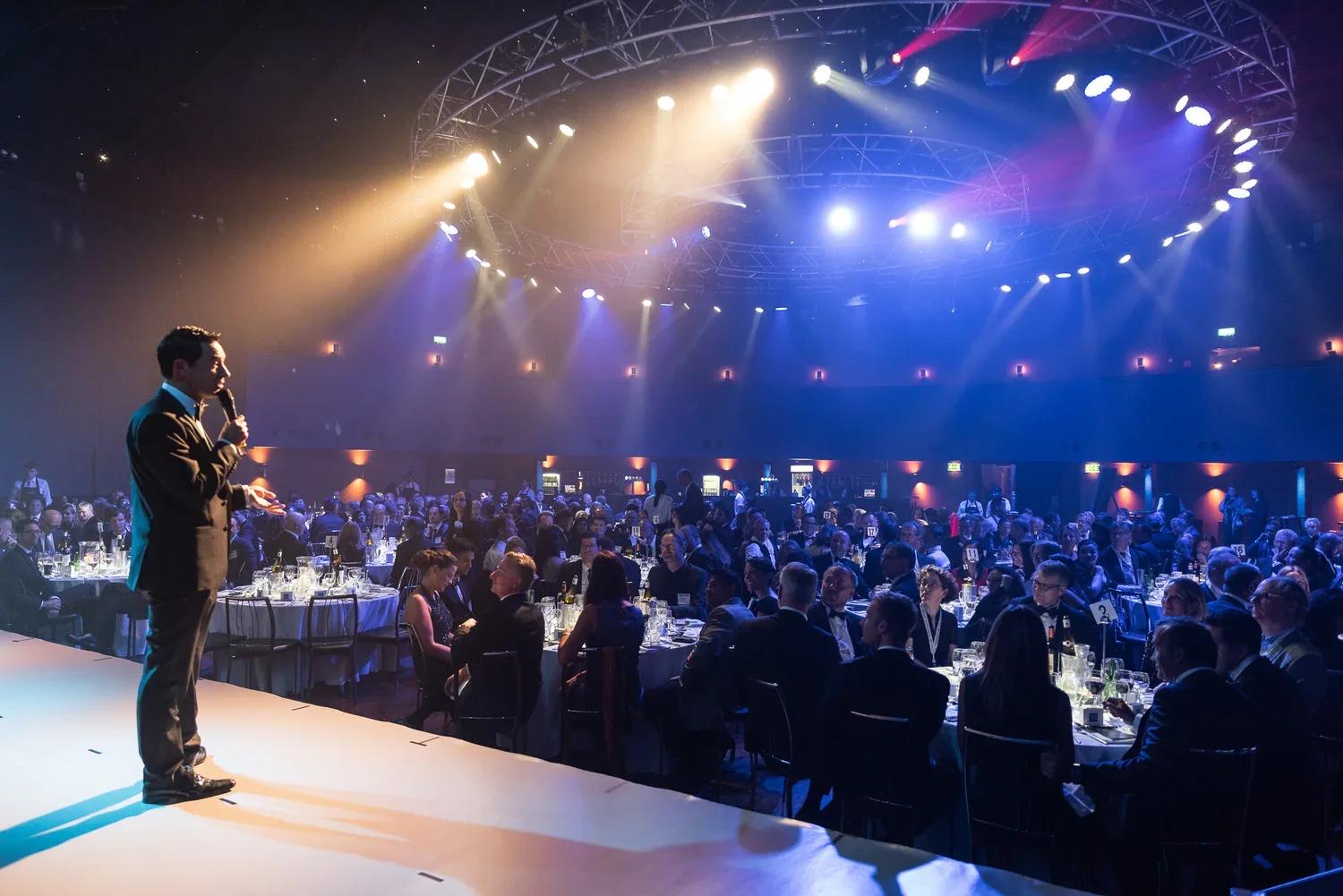 A man in a tuxedo speaking into a microphone on a stage at a formal event, with many guests seated at round tables in a large, dimly lit hall with colorful stage lighting.