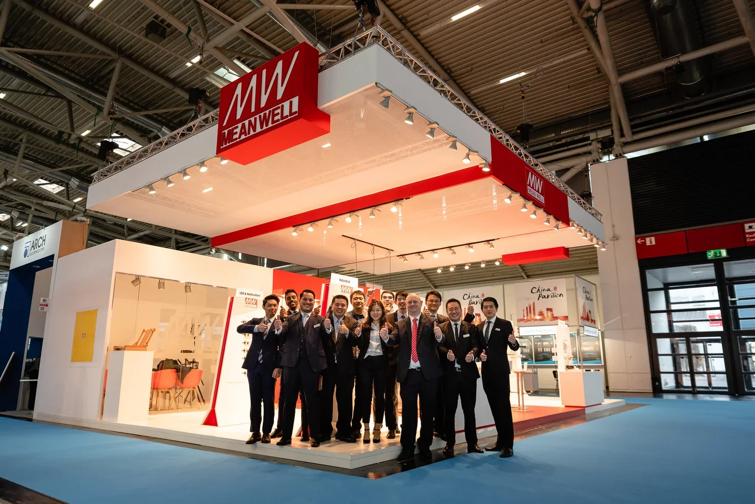Group of business professionals in suits standing in front of a MEANWELL booth at a trade show, giving thumbs up.