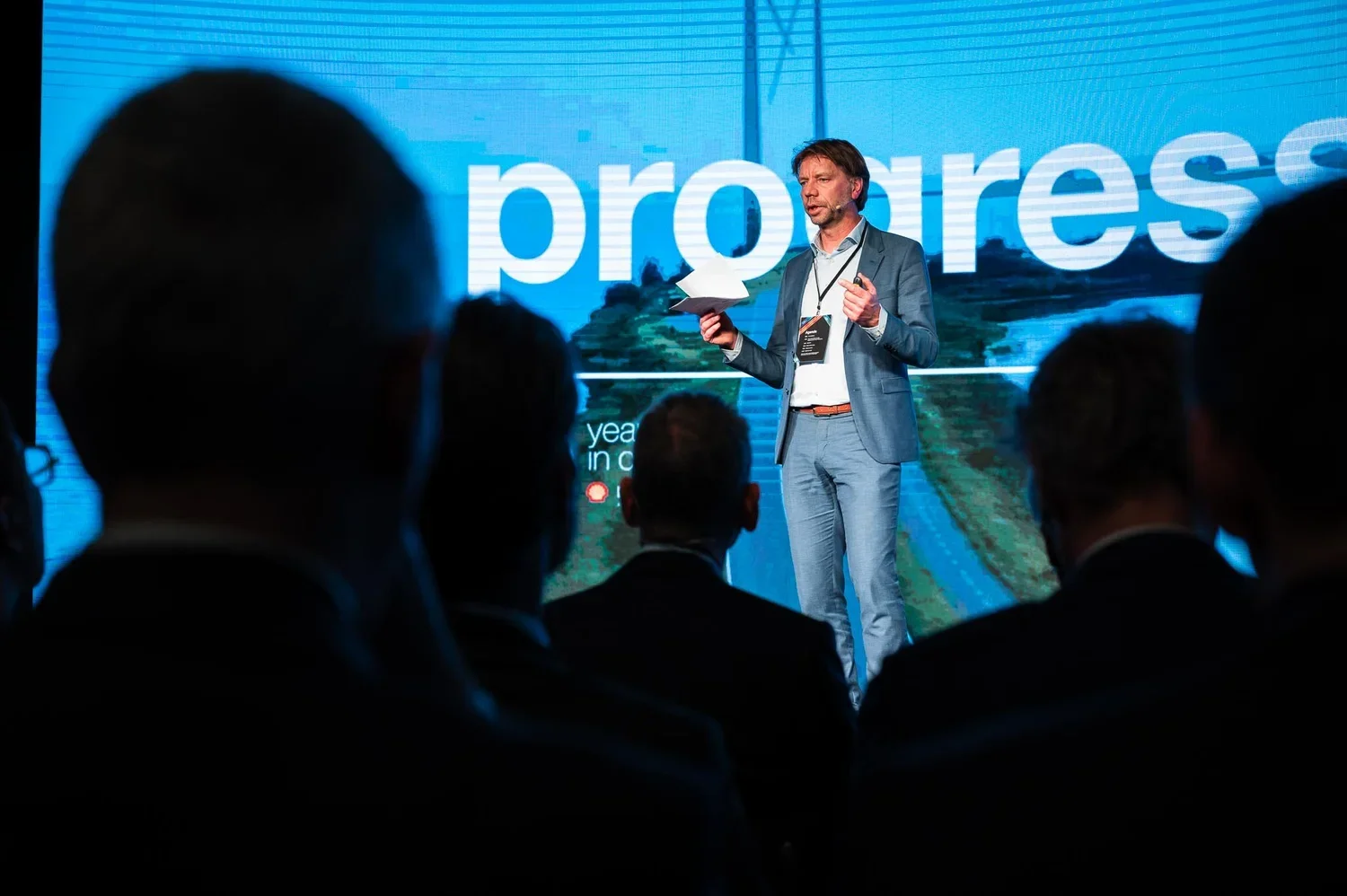 A man in a blue suit giving a presentation on stage at a conference, with a digital screen behind him displaying the word 'progress' and an image of a landscape.