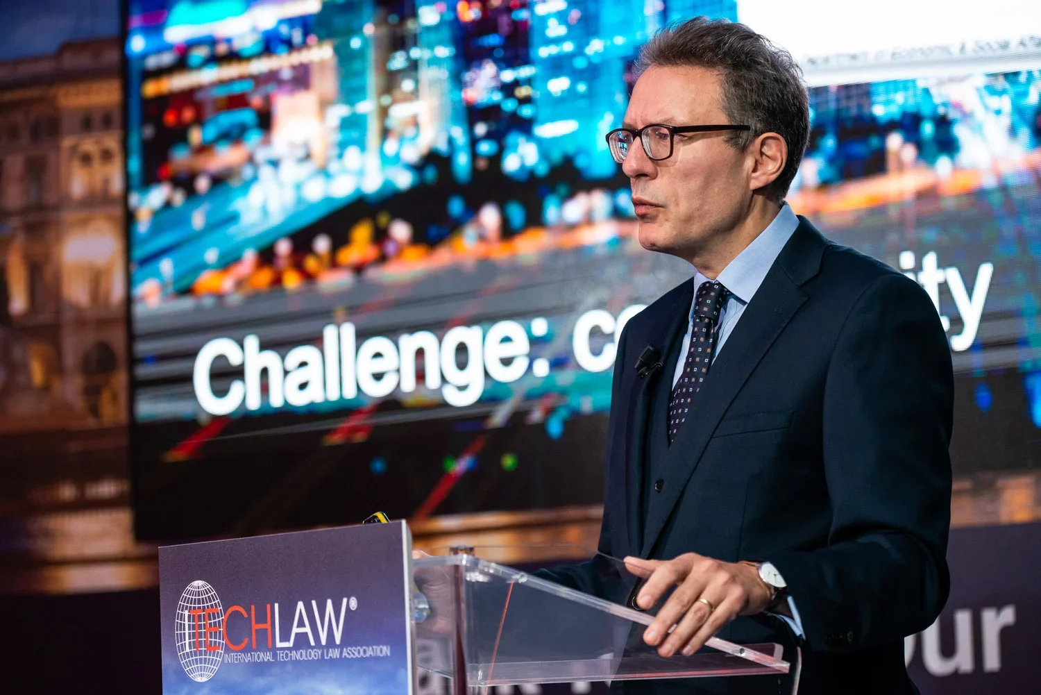 A man in a dark suit and glasses speaking at a podium with a sign reading 'TECHLAw' in front of a large screen displaying the words 'Challenge: c'.