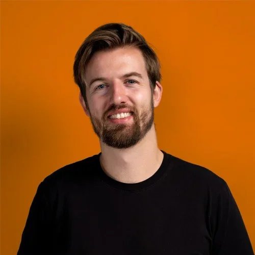 A young man with a beard and mustache, smiling, wearing a black shirt, against an orange background.