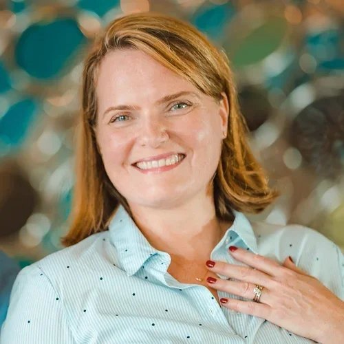 A woman with red hair and a light blue shirt smiling and holding her hand with a wedding ring on her chest.