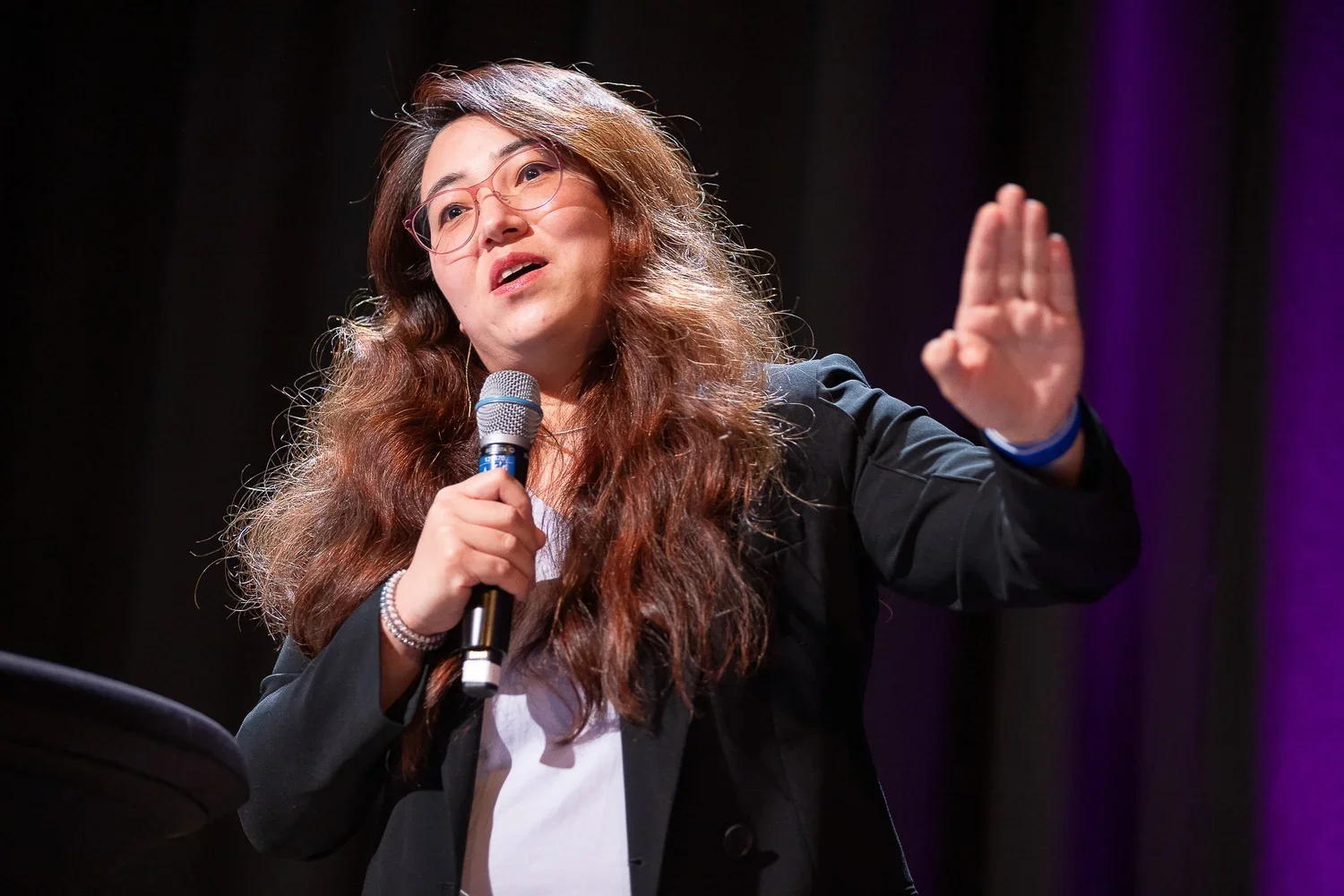 A woman with long brown hair, glasses, and a black blazer speaking into a microphone on stage, gesturing with her right hand.