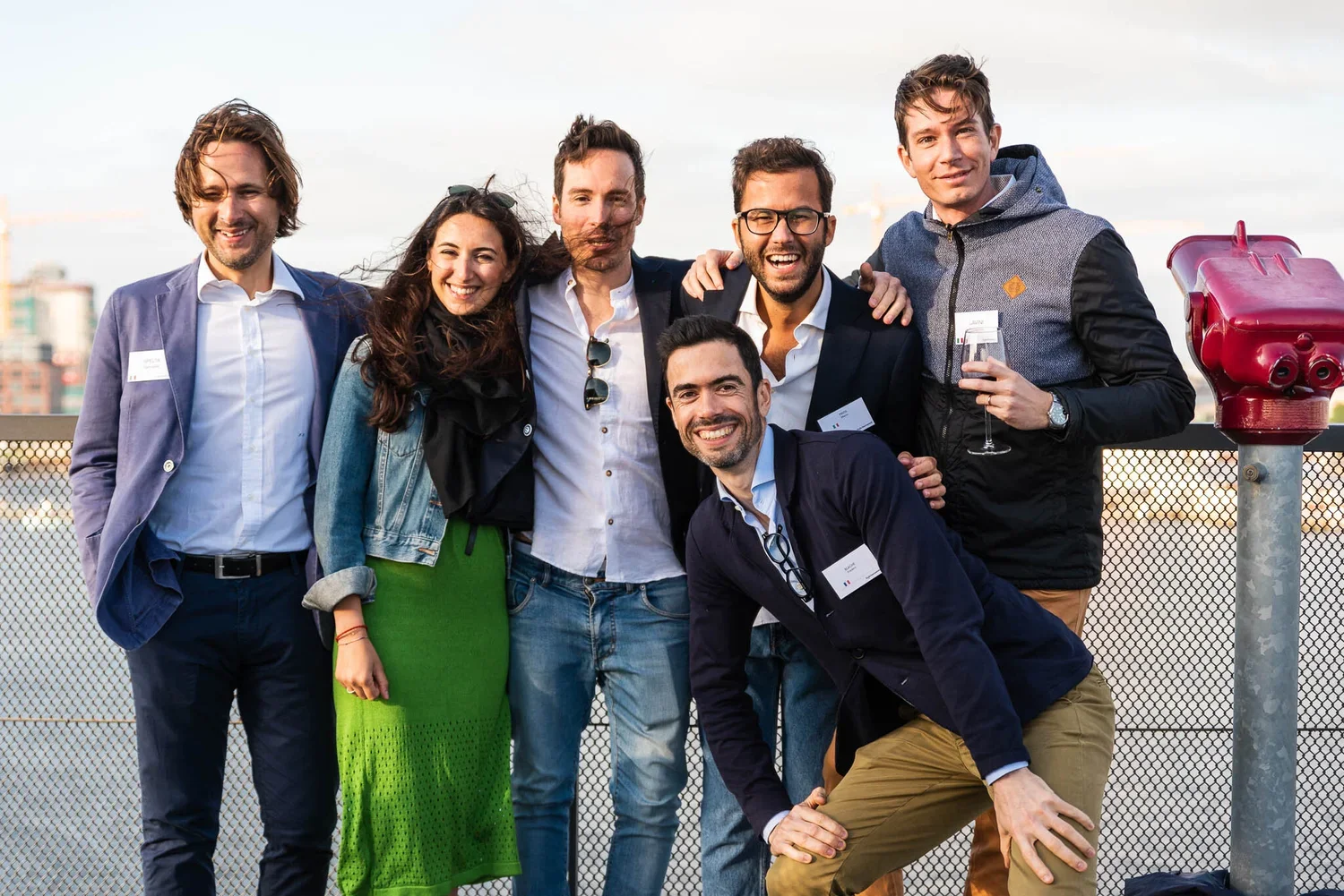 Group of seven young adults smiling and posing together outdoors on a balcony or rooftop, with city buildings in the background.