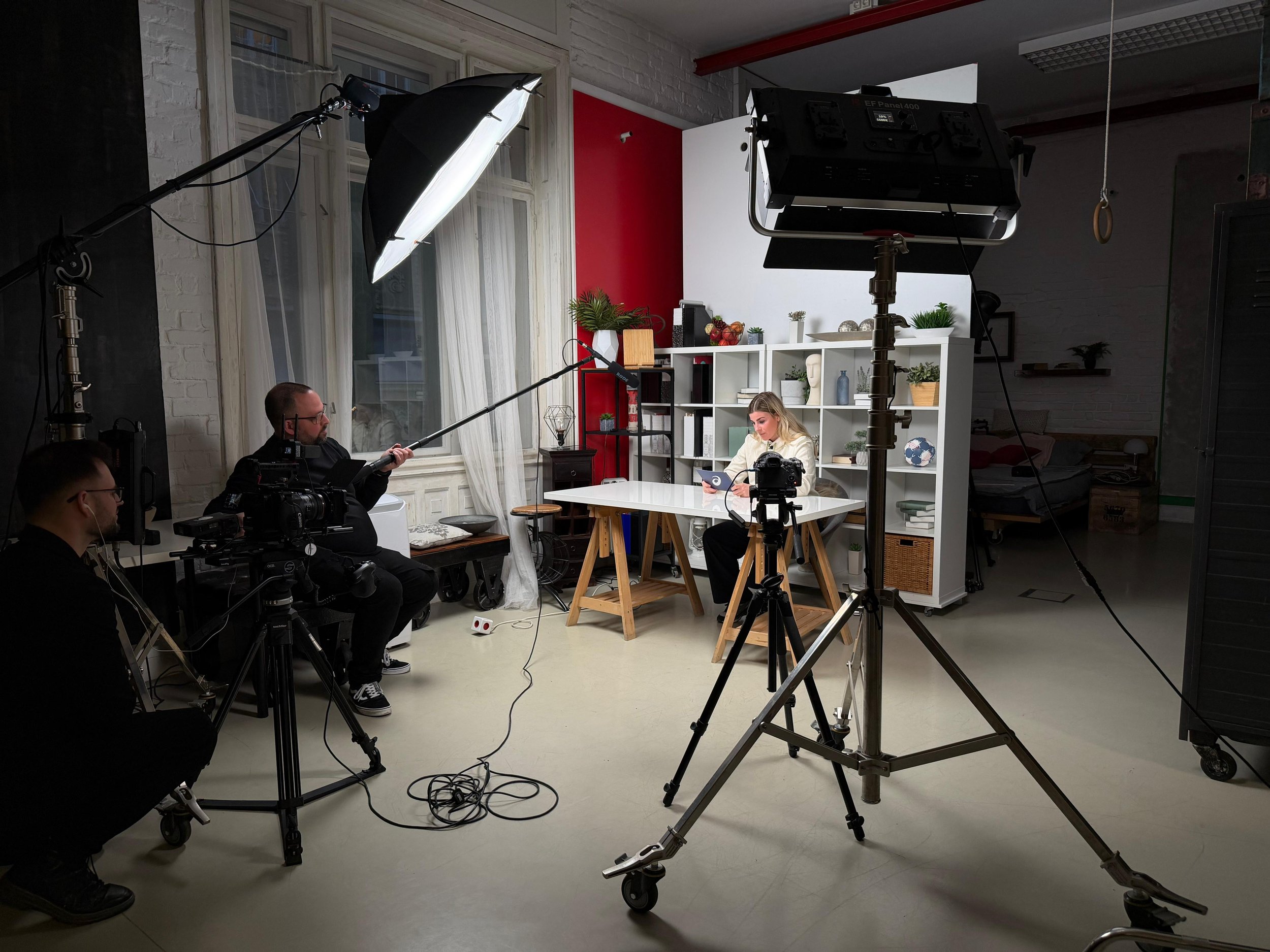 A woman sits at a white desk in a studio with filming equipment, including cameras, lights, and a boom microphone, as she looks at her phone. The studio has white brick walls, large windows, white curtains, and industrial-style decor.