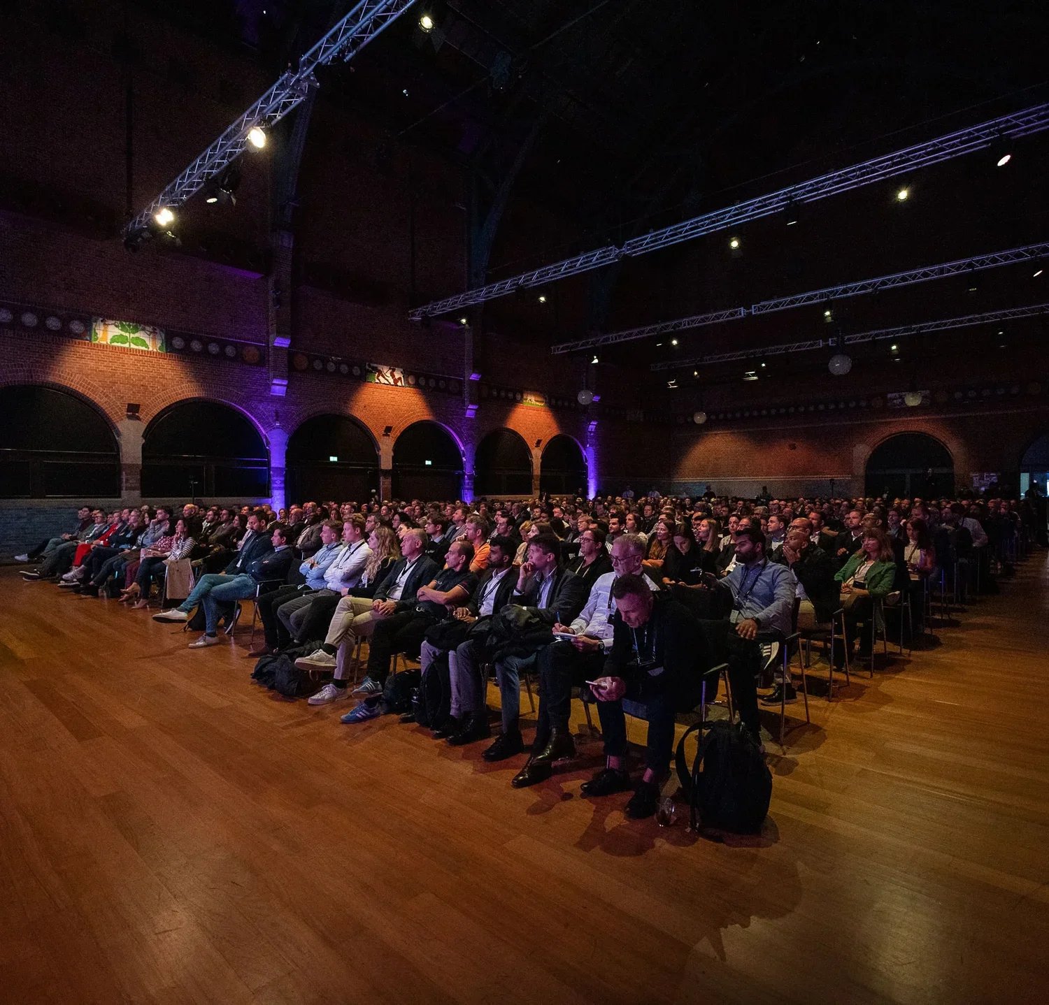 A large audience seated in a conference hall with brick walls and arched windows, attending a presentation or event at night.