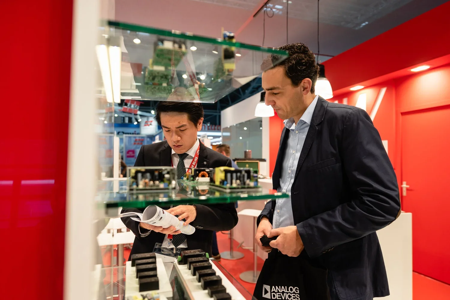 Two men in business attire examining electronic circuit boards and components at a trade show or exhibition, with a red background and display cases.