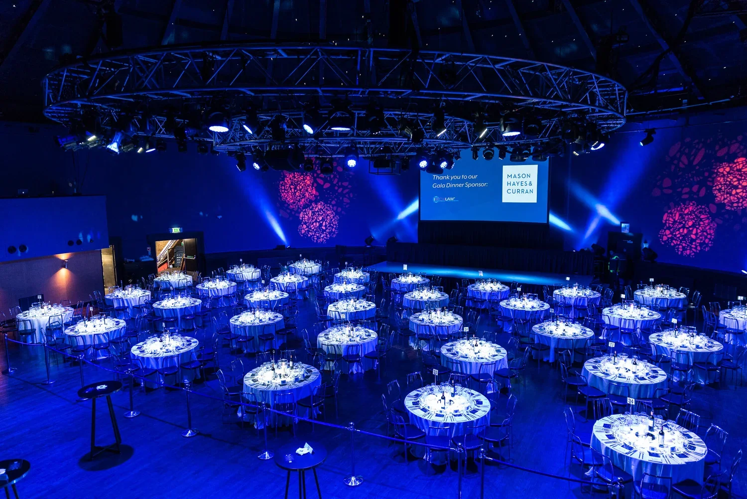 Empty banquet hall with round tables covered in white tablecloths, set for a formal event with wine bottles and glasses, illuminated with blue and purple lighting, a stage with a screen displaying sponsor information, and decorative wall projections.