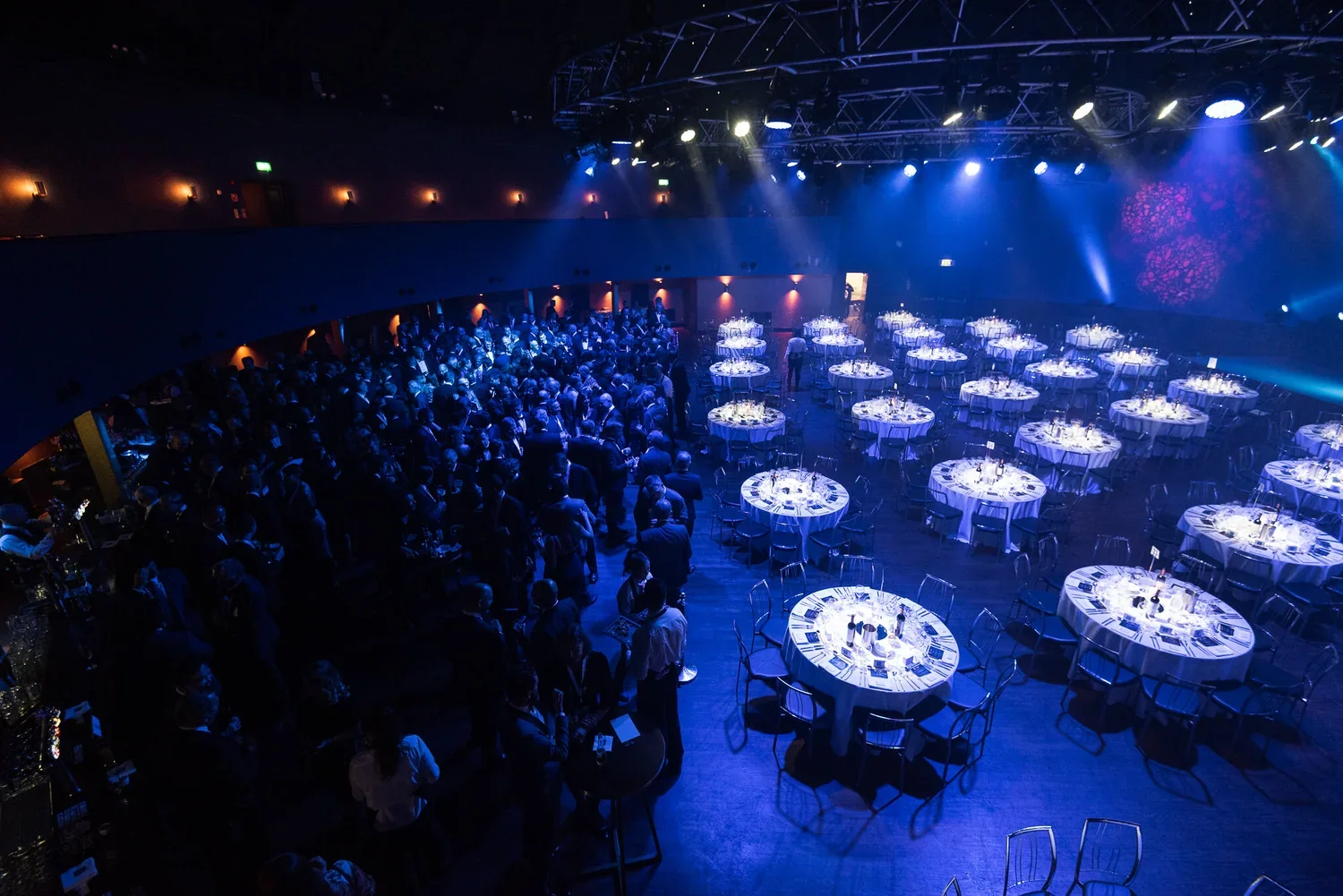 A large banquet hall filled with guests standing and mingling near illuminated round tables with white tablecloths set for dining, under blue stage lighting.
