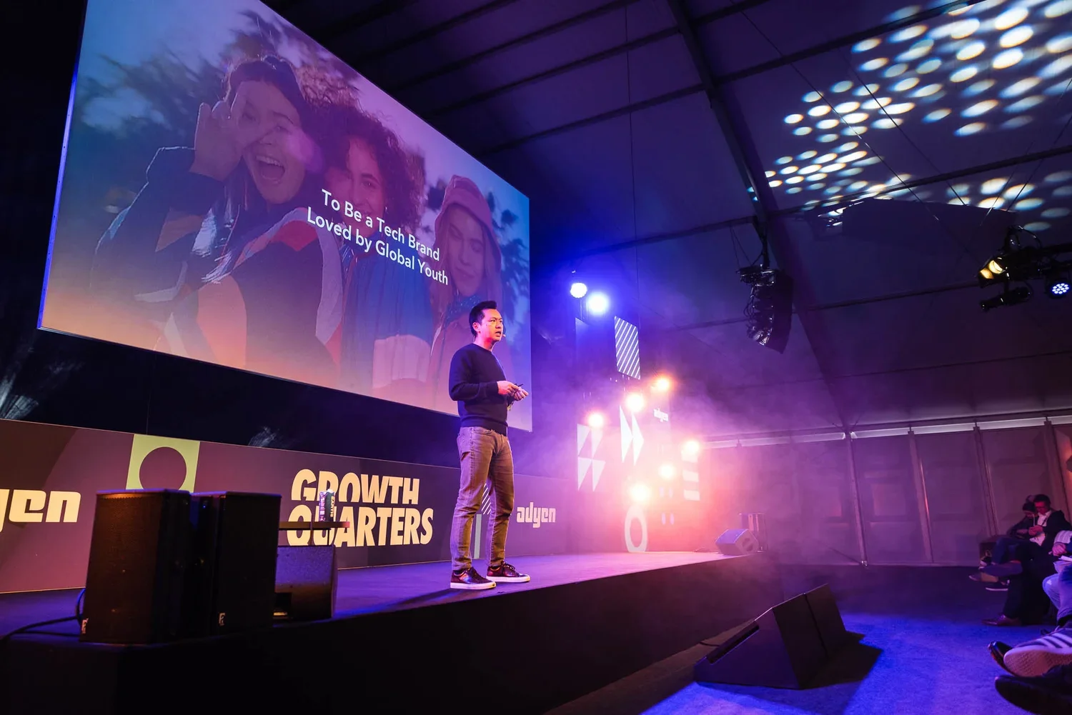 A man standing on stage delivering a presentation at a conference. There is a large screen behind him displaying a slide that reads 'To Be a Tech Brand Loved by Global Youth' with a picture of diverse young people. The stage has bright purple and ora