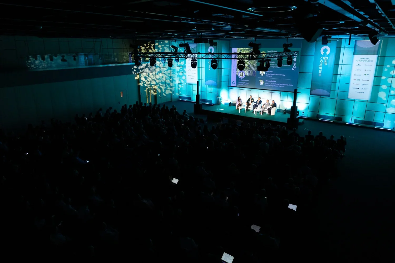 A large conference room hosting a panel discussion with five speakers on stage, illuminated by blue lighting, with a big audience watching from seats, and large screens displaying information behind the panel.