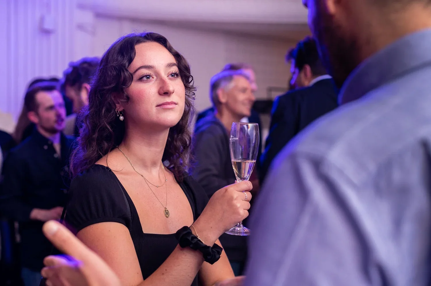 A woman with dark wavy hair holding a glass of champagne in her right hand during a social event, facing a man in a gray shirt. Several other people are in the background, some smiling and talking, in an indoor setting with purple and blue lighting.