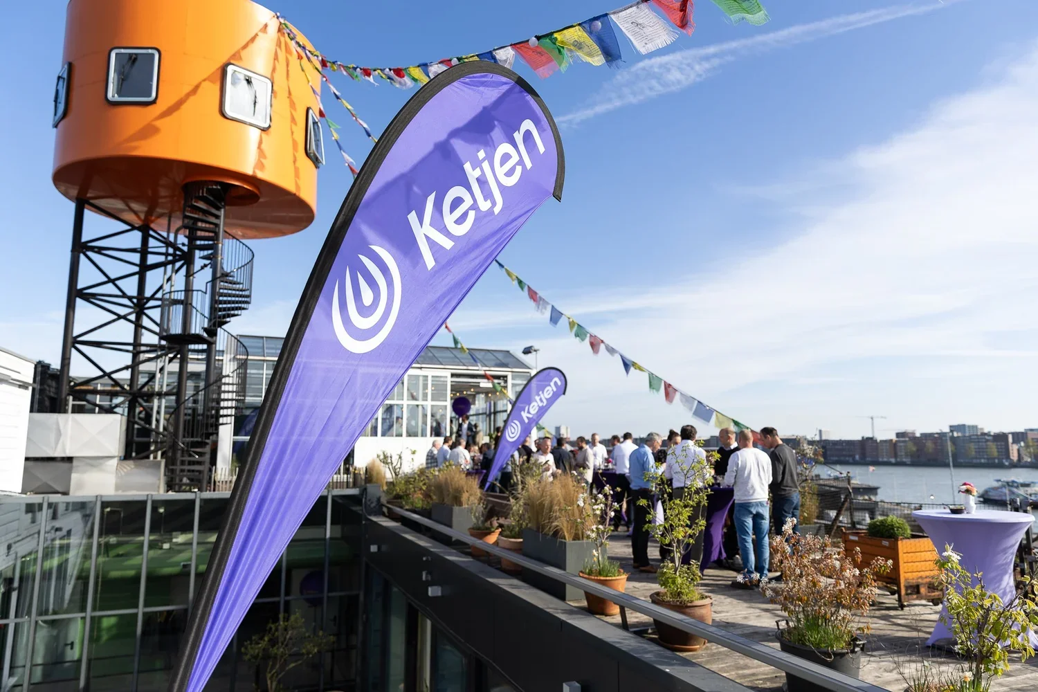 Outdoor rooftop event with people gathered around tables, naval flags, and purple banners displaying the word 'Ketjen'. A large orange structure and a water body are visible in the background under a blue sky with clouds.