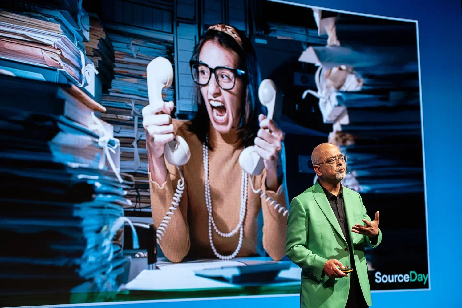 A woman in an office environment appears angry, holding a beige telephone with both hands, with stacks of papers and files around her. In the foreground, a man in a green blazer is speaking, holding a microphone, in front of a large screen displaying