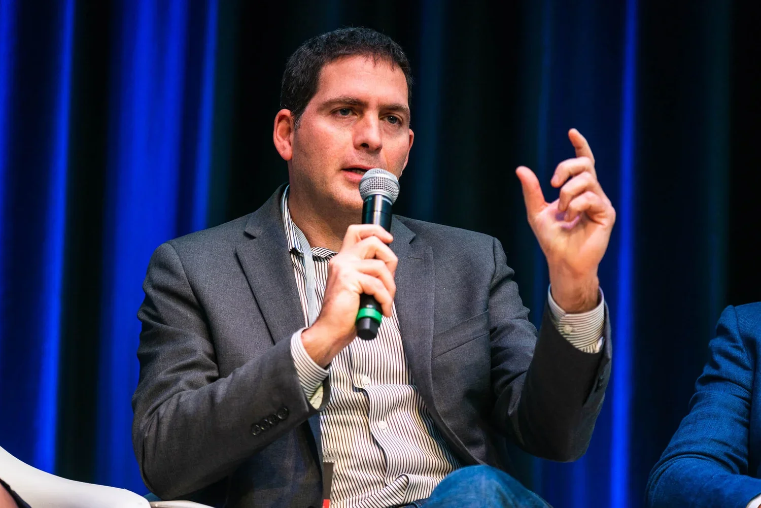 A man in a gray suit holding a microphone and speaking during a panel discussion, with dark curtain backdrop.