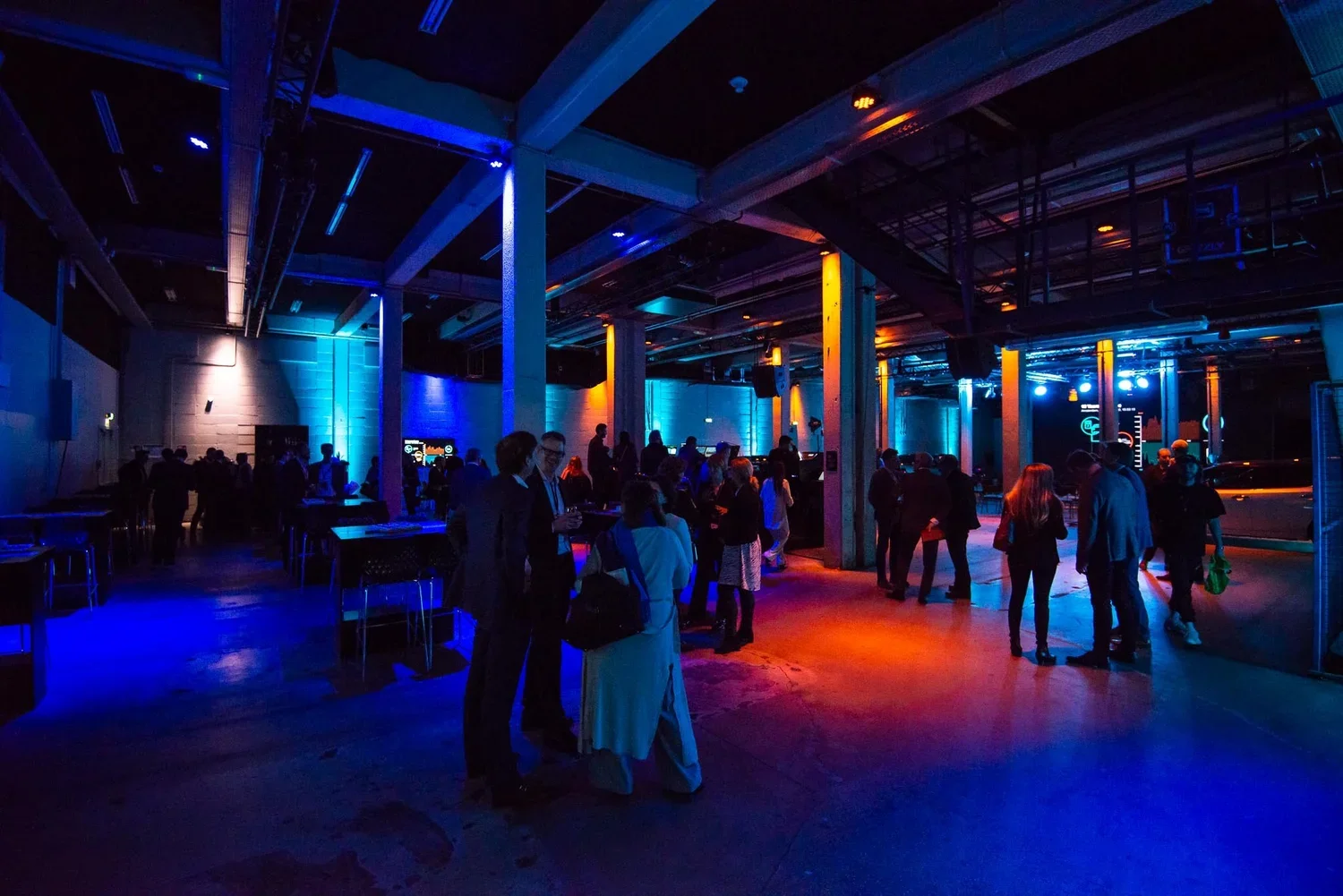 People gathered in a dimly lit industrial-style event space with colorful blue and orange lighting, some standing and socializing.