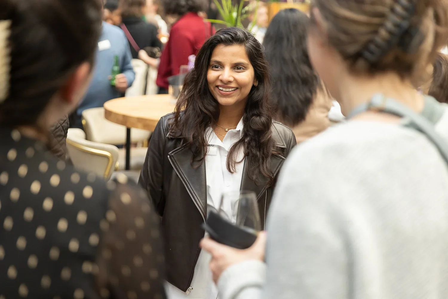 A woman with long dark hair smiling while talking to a group of people at a social gathering in a bright, lively setting.