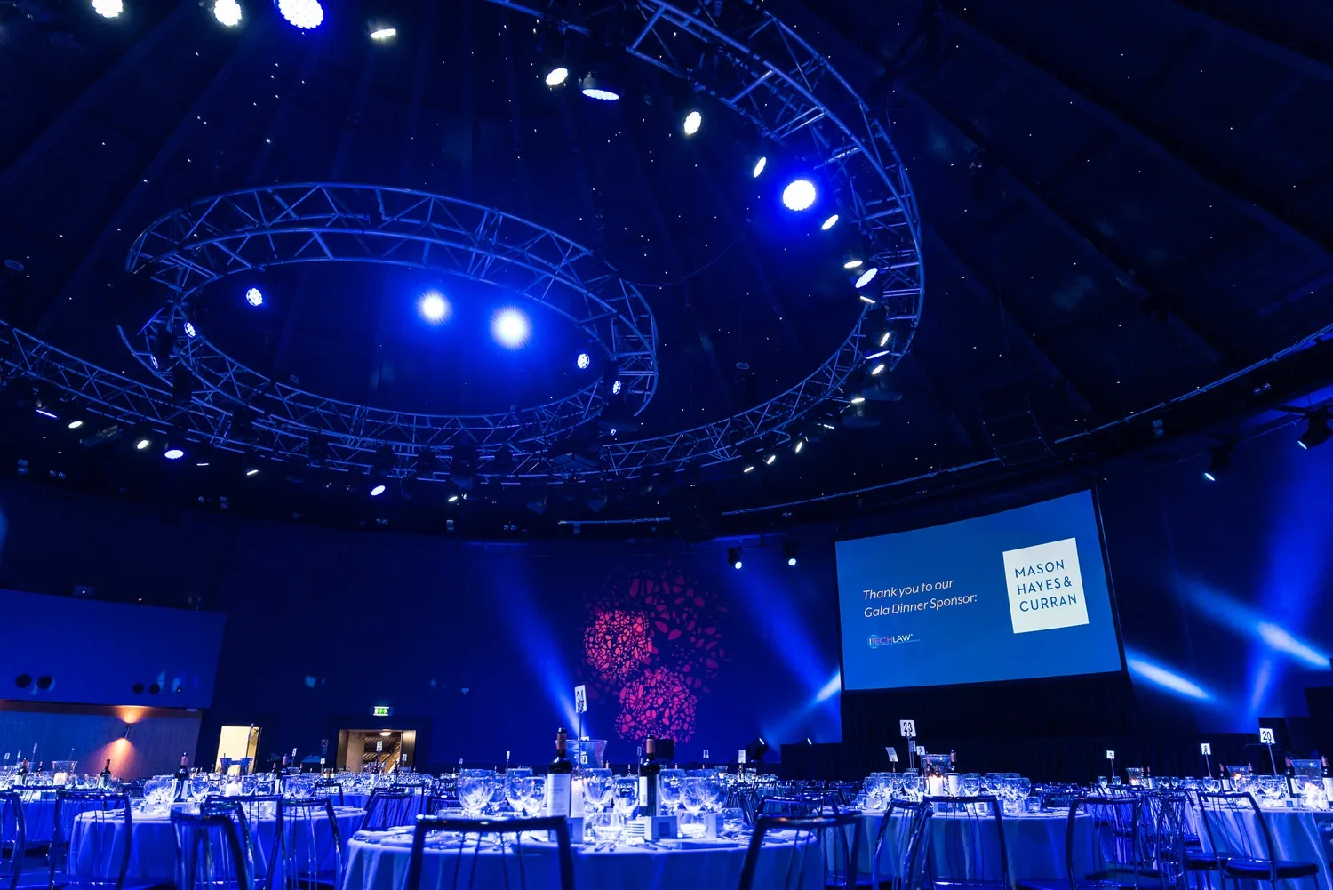 Upscale event hall decorated with round tables set for a formal dinner, illuminated by blue lighting, with a large screen displaying a thank you message to Gala Dinner Sponsor Mason Hayes & Curran and Techlaw, and circular lighting fixtures hanging f