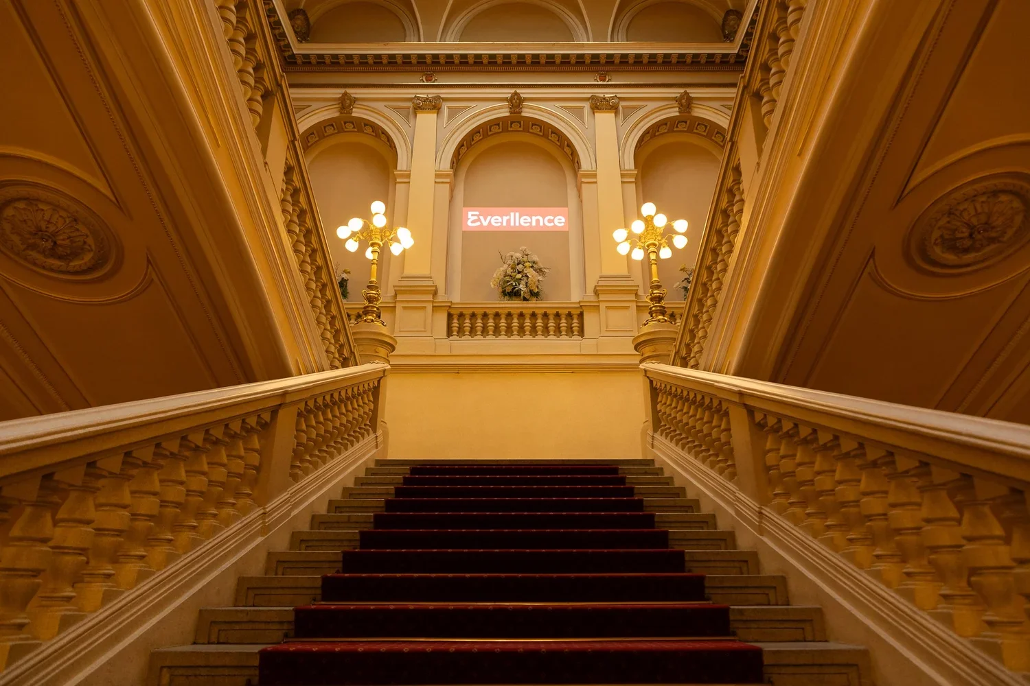Elegant staircase in a grand building with ornate walls and a floral arrangement at the top, lit by chandelier-style lamps, with a sign that reads 'Everllence'.