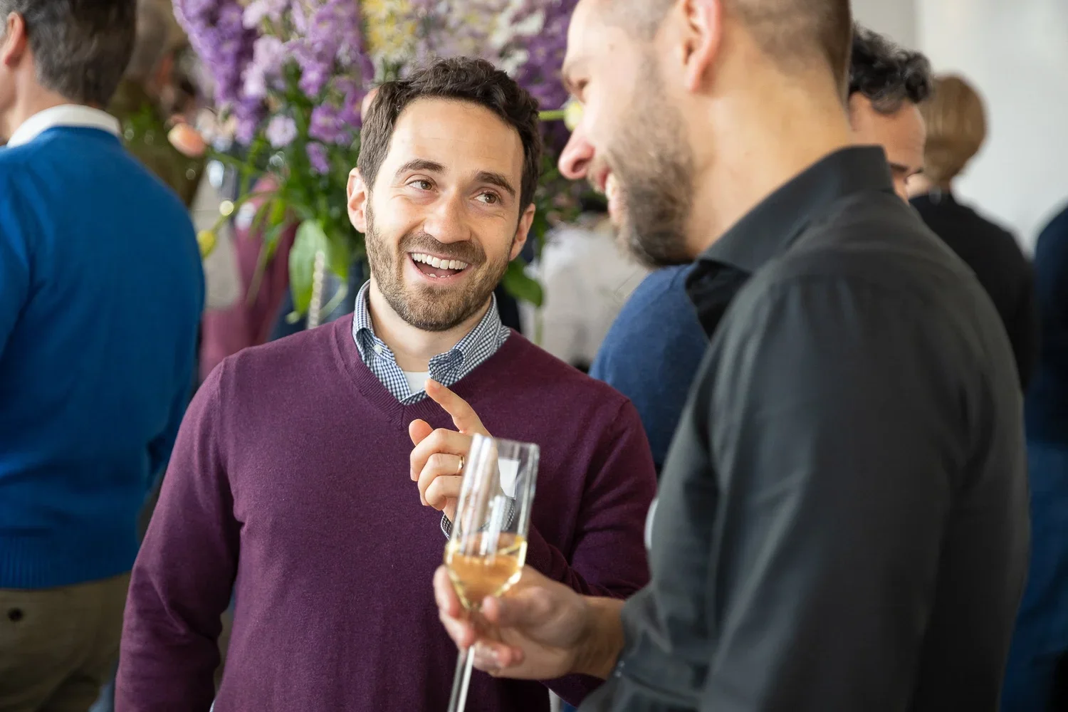 Two men are chatting at a social gathering; one is laughing while holding a champagne flute, and the other is smiling and gesturing with his finger in a friendly manner. The background features other people and purple flowers.