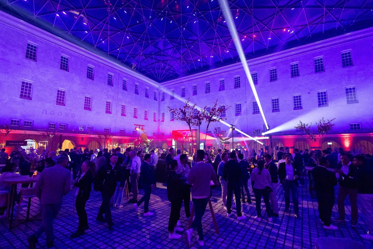 A lively indoor event with people mingling under purple and blue lighting, with trees and beams of light projecting onto a high ceiling.