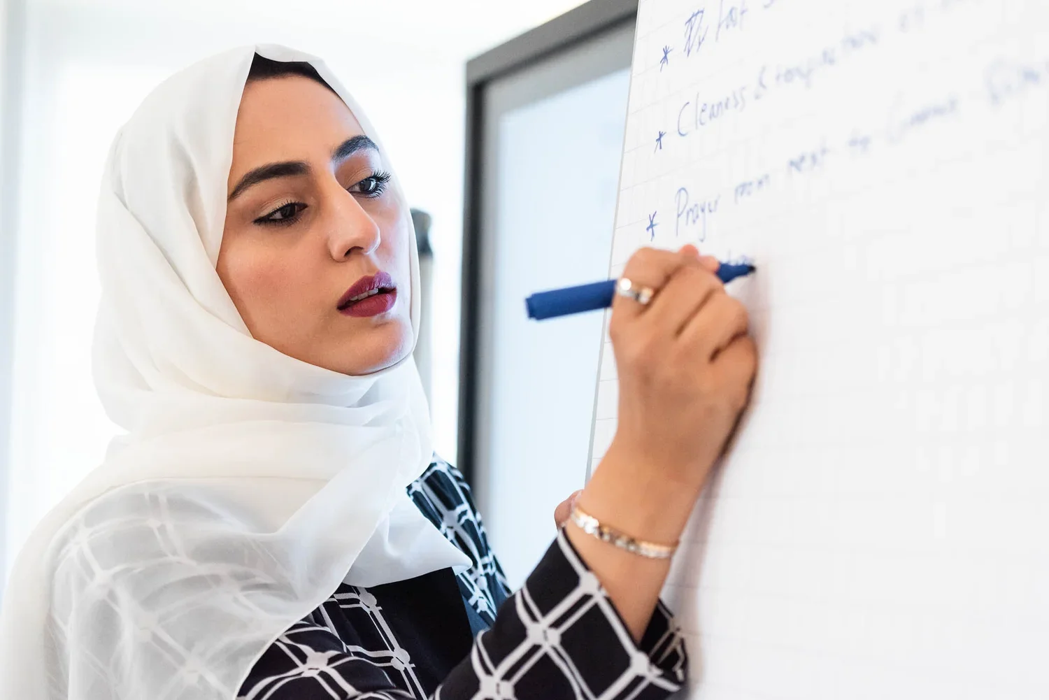 Woman in a white headscarf writing on a whiteboard. She appears focused and is wearing a black and white patterned outfit.