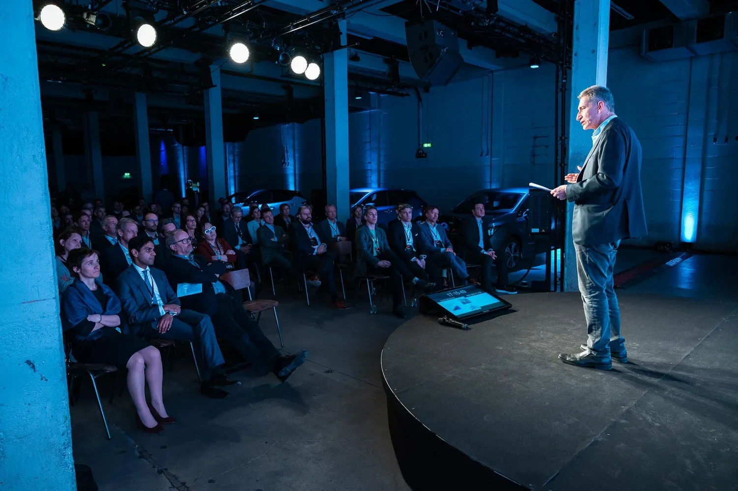 A man giving a presentation on a stage to an audience seated in a dark, industrial-style venue with cars in the background and stage lights overhead.